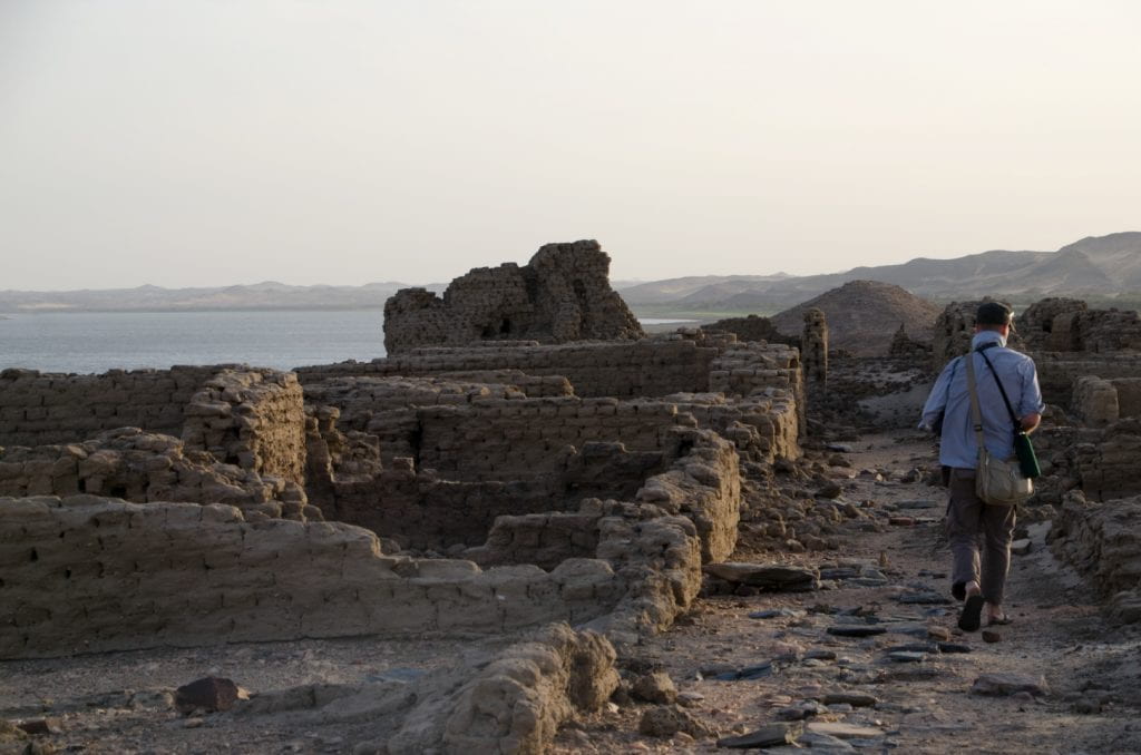 photo of man walking down ancient street away from camera, ruins of mudbrick walls on either side