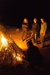 photo of four people standing near a fire of thin sticks at night